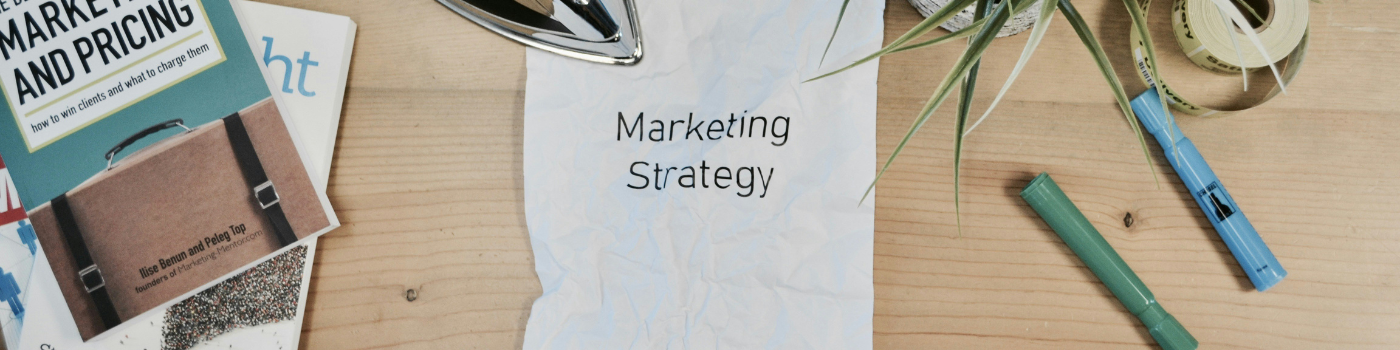 Vue de dessus d'un bureau en bois comprenant un livre intitulé "Marketing and Pricing", une feuille froissée au centre portant l'inscription "Marketing Strategy", un fer à repasser, des feutres et une plante verte.