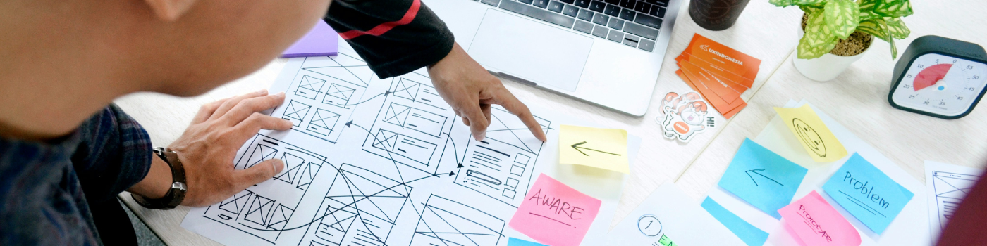 Vue de dessus d'un designer travaillant sur des croquis de wireframes avec des post-it colorés illustrant les étapes du Design Thinking : Empathize, Define et Ideate.