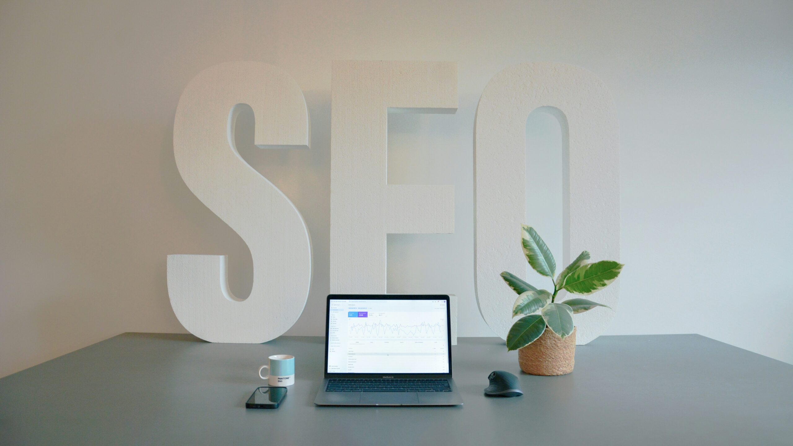 Un ordinateur portable ouvert affichant des graphiques d'analyse de données sur un bureau gris, devant de grandes lettres blanches formant le mot SEO, une tasse de café, un smartphone et une plante verte en pot.
