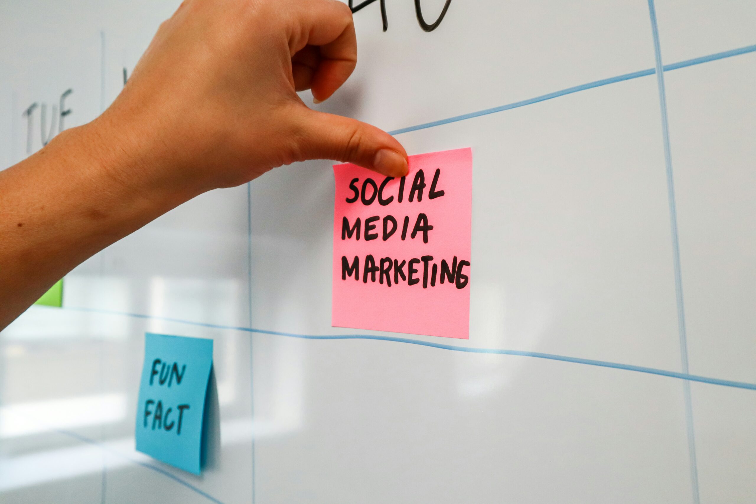 Une main place une note autocollante rose portant l'inscription « SOCIAL MEDIA MARKETING » sur un tableau blanc de planification.
