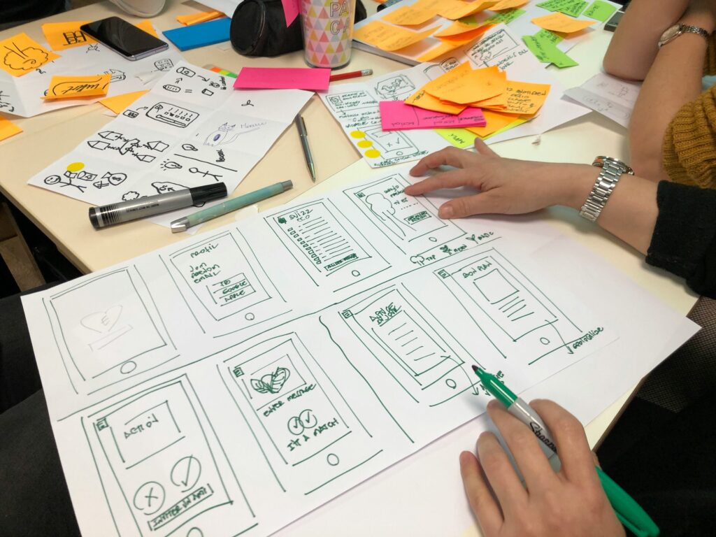 Vue en plongée d'une séance de remue-méninges et de conception UX/UI. Deux personnes travaillent autour d'une grande table encombrée. Elles dessinent des maquettes d'applications mobiles (wireframes) sur une grande feuille de papier blanc à l'aide de feutres. La table est parsemée de nombreuses notes adhésives colorées (post-it) jaunes et roses, de stylos, de crayons et de feuilles de brouillon affichant des croquis d'icônes et de symboles graphiques.