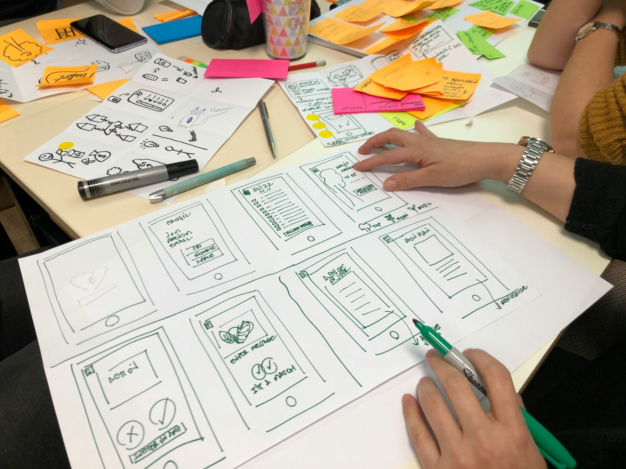 Vue en plongée d'une séance de remue-méninges et de conception UX/UI. Deux personnes travaillent autour d'une grande table encombrée. Elles dessinent des maquettes d'applications mobiles (wireframes) sur une grande feuille de papier blanc à l'aide de feutres. La table est parsemée de nombreuses notes adhésives colorées (post-it) jaunes et roses, de stylos, de crayons et de feuilles de brouillon affichant des croquis d'icônes et de symboles graphiques.